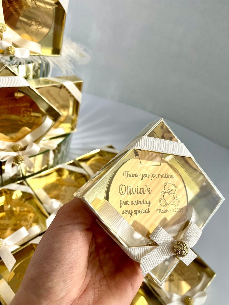 May include: Gold favor boxes with a clear lid and a gold mirror inside. The mirror has a personalized message that says "Thank you for making Olivia's first birthday very special" with a teddy bear graphic and the date "March 30, 2023".