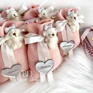 May include: Pink fabric gift bags with white ribbon and a small white teddy bear with a silver bow. Each bag has a silver heart-shaped tag with the name "Lamita" written on it.