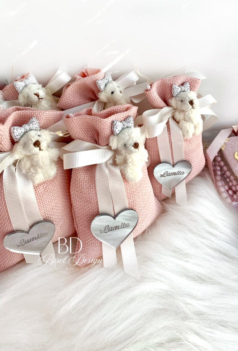 May include: Pink fabric gift bags with white ribbon and a small white teddy bear with a silver bow. Each bag has a silver heart-shaped tag with the name "Lamita" written on it.