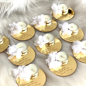May include: Gold round favor boxes with white flowers and tea lights. Each box has a personalized message that says "Merci de partager ce moment avec nous. Iris & Hamza. 23.10.2021".