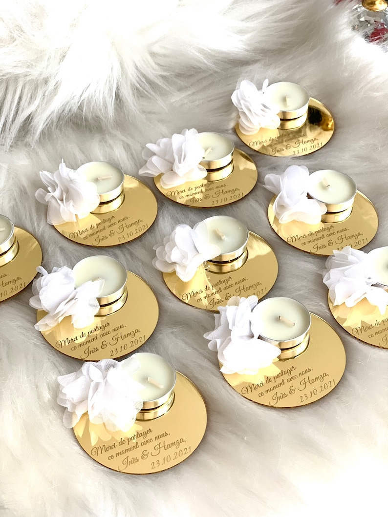 May include: Gold round favor boxes with white flowers and tea lights. Each box has a personalized message that says "Merci de partager ce moment avec nous. Iris & Hamza. 23.10.2021".