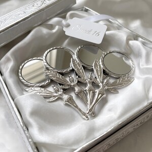 May include: A set of silver-toned metal hand mirrors, each featuring a fairy figure with detailed wings as the handle. The mirrors are arranged inside a white satin-lined box with a white tag attached that reads Sweet 16 in cursive script.