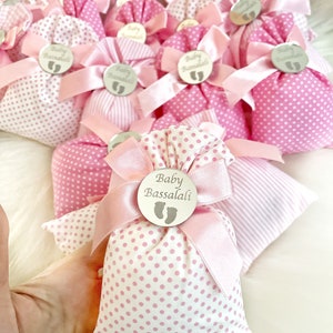 May include: Pink and white polka dot fabric gift bags with pink ribbon bows and silver circular tags that say "Baby Bassalali" with a baby footprint symbol.