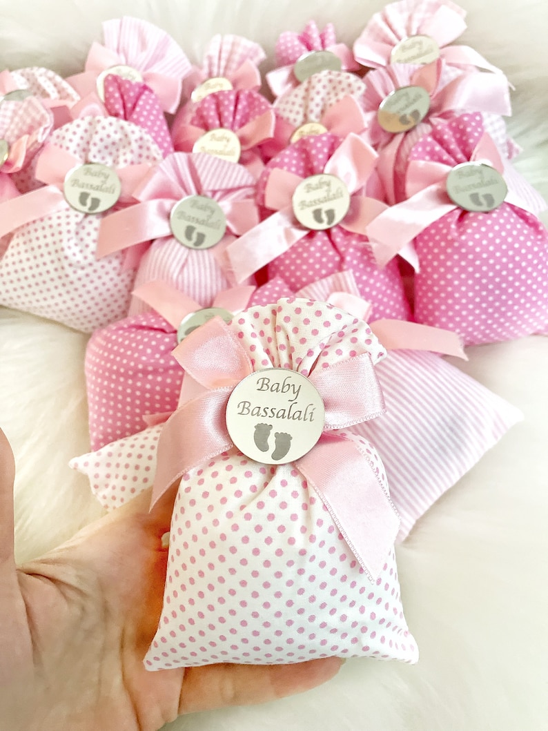 May include: Pink and white polka dot fabric gift bags with pink ribbon bows and silver circular tags that say "Baby Bassalali" with a baby footprint symbol.