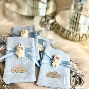 Puede incluir: Bolsitas de regalo azul claro con un encanto de oso de peluche blanco y una etiqueta plateada con el nombre "Vian" y la fecha "01-10-2020".