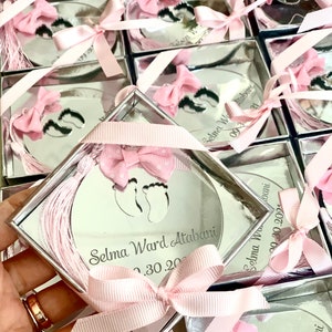 Op de afbeelding: Een ronde, gespiegelde herinneringsdoos van zilver met een roze lint en strik. De doos heeft een uitsnijding van twee babyvoetjes en de tekst "Selma Ward Atabani 02.30.2024".
