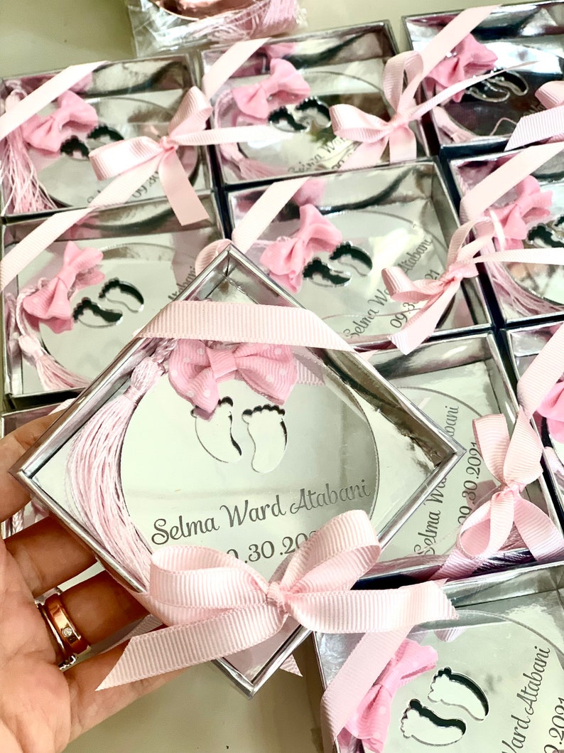Op de afbeelding: Een ronde, gespiegelde herinneringsdoos van zilver met een roze lint en strik. De doos heeft een uitsnijding van twee babyvoetjes en de tekst "Selma Ward Atabani 02.30.2024".
