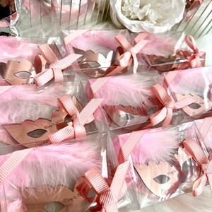 May include: Pink and gold party favours in clear boxes with pink feathers and a pink ribbon. Each favour features a rose gold mirror mask with the text "Sienna's" and "May 6, 2023".