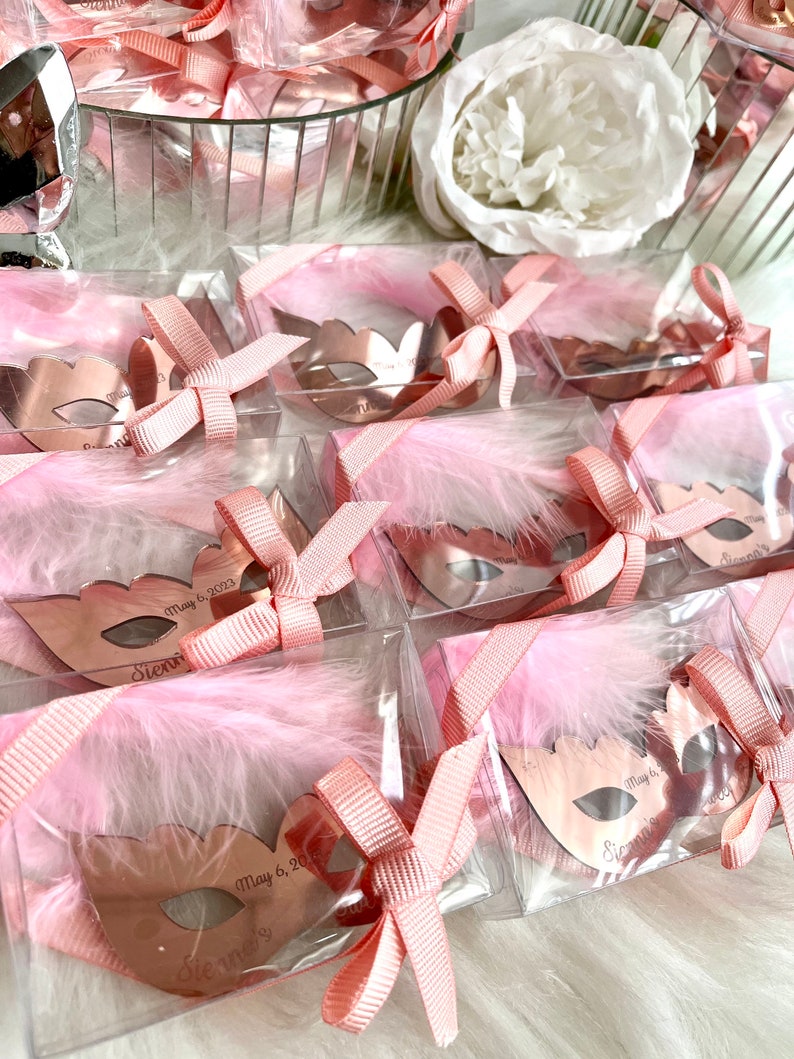 May include: Pink and gold party favours in clear boxes with pink feathers and a pink ribbon. Each favour features a rose gold mirror mask with the text "Sienna's" and "May 6, 2023".
