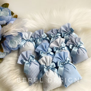 May include: A group of small blue and white fabric gift bags with blue ribbon bows. The bags are filled with a white material and are arranged on a white fluffy surface.
