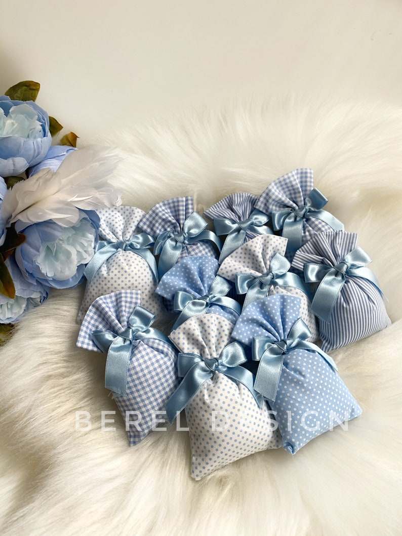 May include: A group of small blue and white fabric gift bags with blue ribbon bows. The bags are filled with a white material and are arranged on a white fluffy surface.