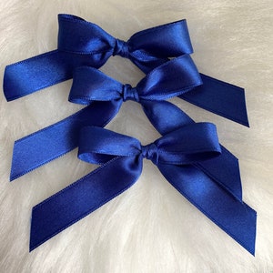 May include: Three blue satin ribbon bows tied together. The bows are arranged in a stack, with the largest bow on the bottom and the smallest bow on top.