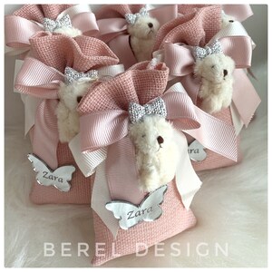 May include: Pink fabric gift bags with white teddy bears, pink bows, and silver butterfly charms with the name "Zara" on them. The bags are tied with white and pink ribbon.