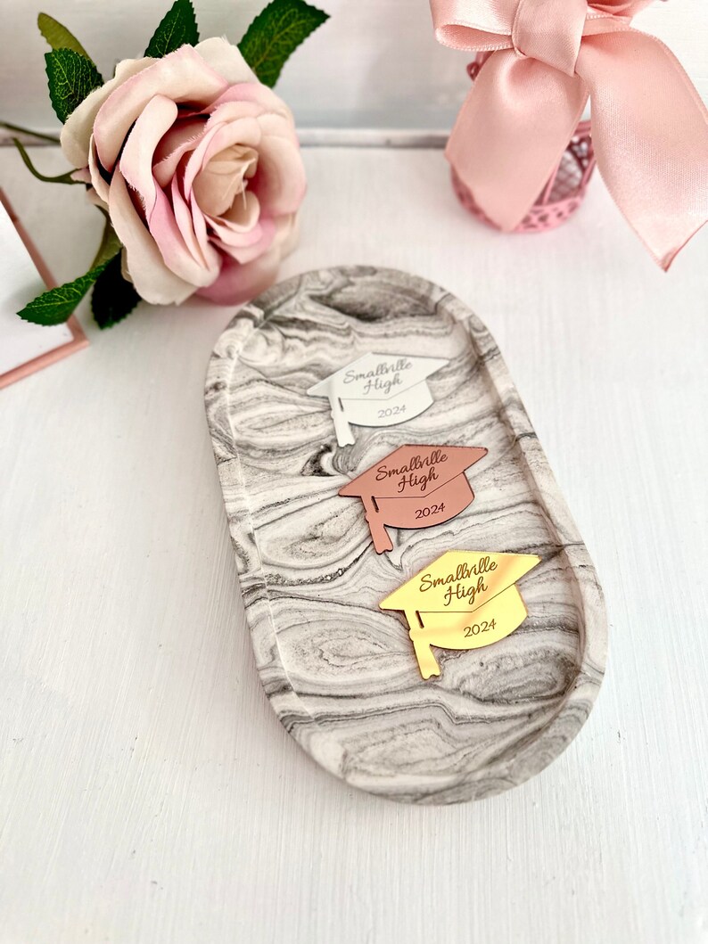 May include: Three graduation caps with the text "Smallville High 2024" on a marble tray. The caps are silver, rose gold, and gold.