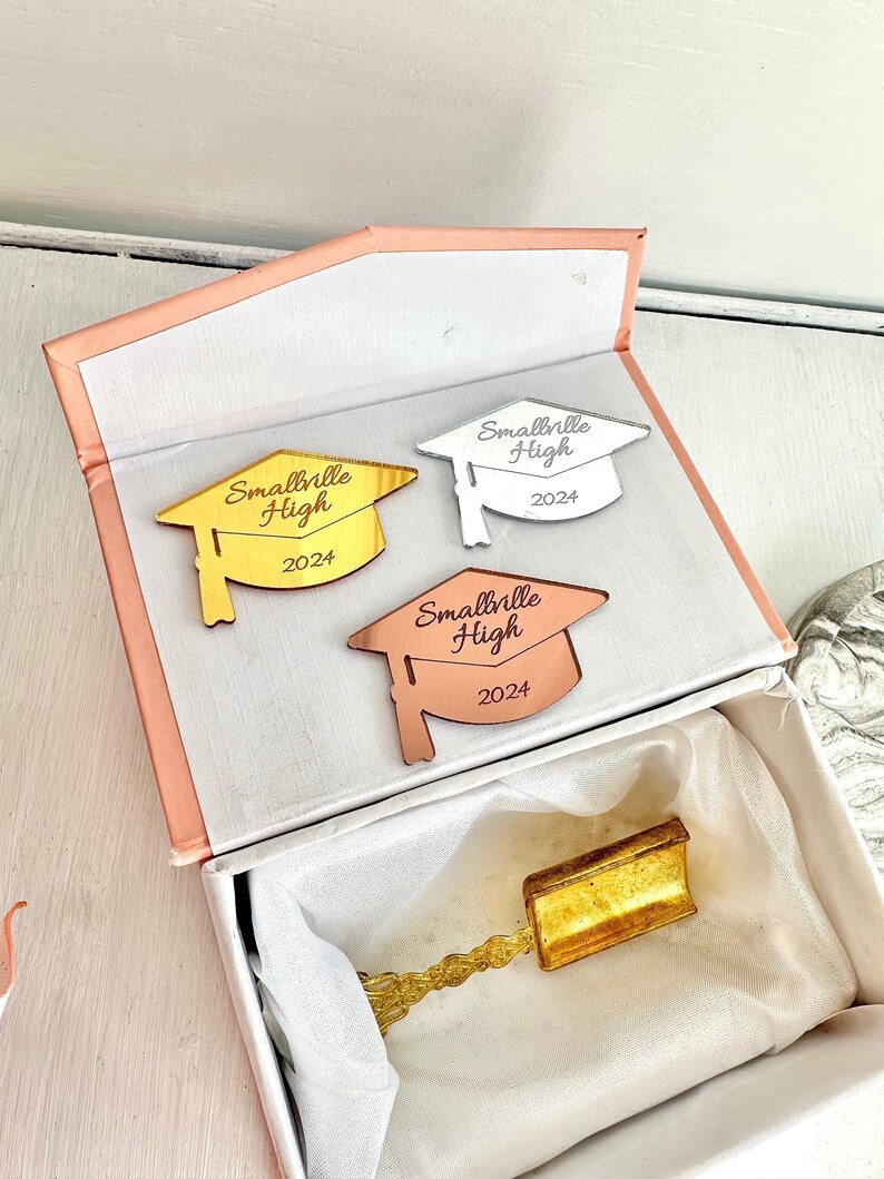 May include: Three gold, silver, and rose gold graduation caps with the text "Smallville High 2024" on each. The caps are in a white box with a pink lid.