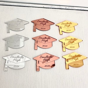 May include: Nine graduation cap-shaped acrylic ornaments in three different colors: silver, rose gold, and gold. Each ornament features the text "Smallville High 2024".