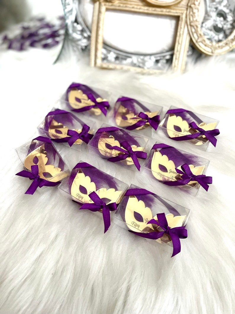 Puede incluir: Una colecci&oacute;n de peque&ntilde;as cajas transparentes con m&aacute;scaras doradas de carnaval en su interior. Cada caja est&aacute; atada con una cinta morada y tiene un nombre escrito en la m&aacute;scara. Las cajas est&aacute;n dispuestas sobre una superficie blanca y esponjosa.