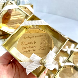 May include: Gold-colored round mirror with a crown and a teddy bear design. The mirror says "Thank you for making Olivia's first birthday very special March 30, 2022". The mirror is in a clear plastic box with a white ribbon.