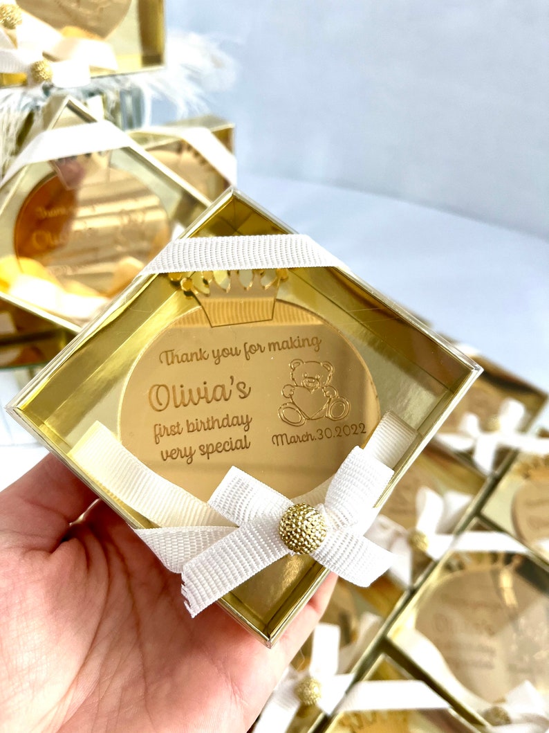 May include: Gold-colored round mirror with a crown and a teddy bear design. The mirror says "Thank you for making Olivia's first birthday very special March 30, 2022". The mirror is in a clear plastic box with a white ribbon.