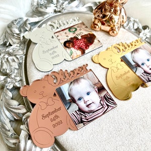 May include: Three personalized mirrored acrylic bear ornaments with a photo frame. The ornaments are in silver, gold, and rose gold. Each ornament has the name "Oliver" and the date "September 24th, 2022".