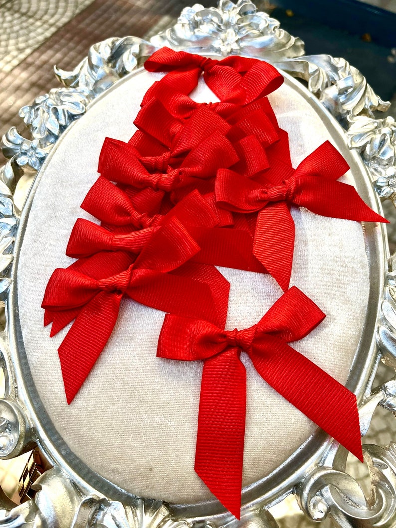 May include: A set of ten red satin ribbon bows, arranged in a decorative pattern on a white background. The bows are tied with a simple knot and have a classic, elegant look.