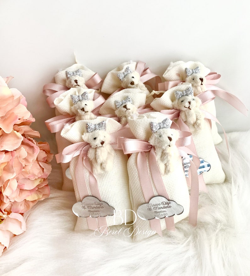 May include: A group of white fabric bags with pink ribbon bows and a small white teddy bear with a silver crown on top. Each bag has a silver cloud-shaped tag with the text "Maeve Eliye B. Mordeno Baptism 14/0" or "Maeve Eliye B. Mordeno Baptism 15/4/20".