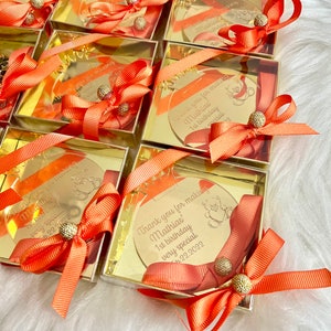 May include: Gold favor boxes with clear lids and orange ribbons. Each box contains a round, gold mirror with a personalized message that says "Thank you for making Mathias' 1st birthday very special." The date "Feb 22, 2022" is also included on the mirror.