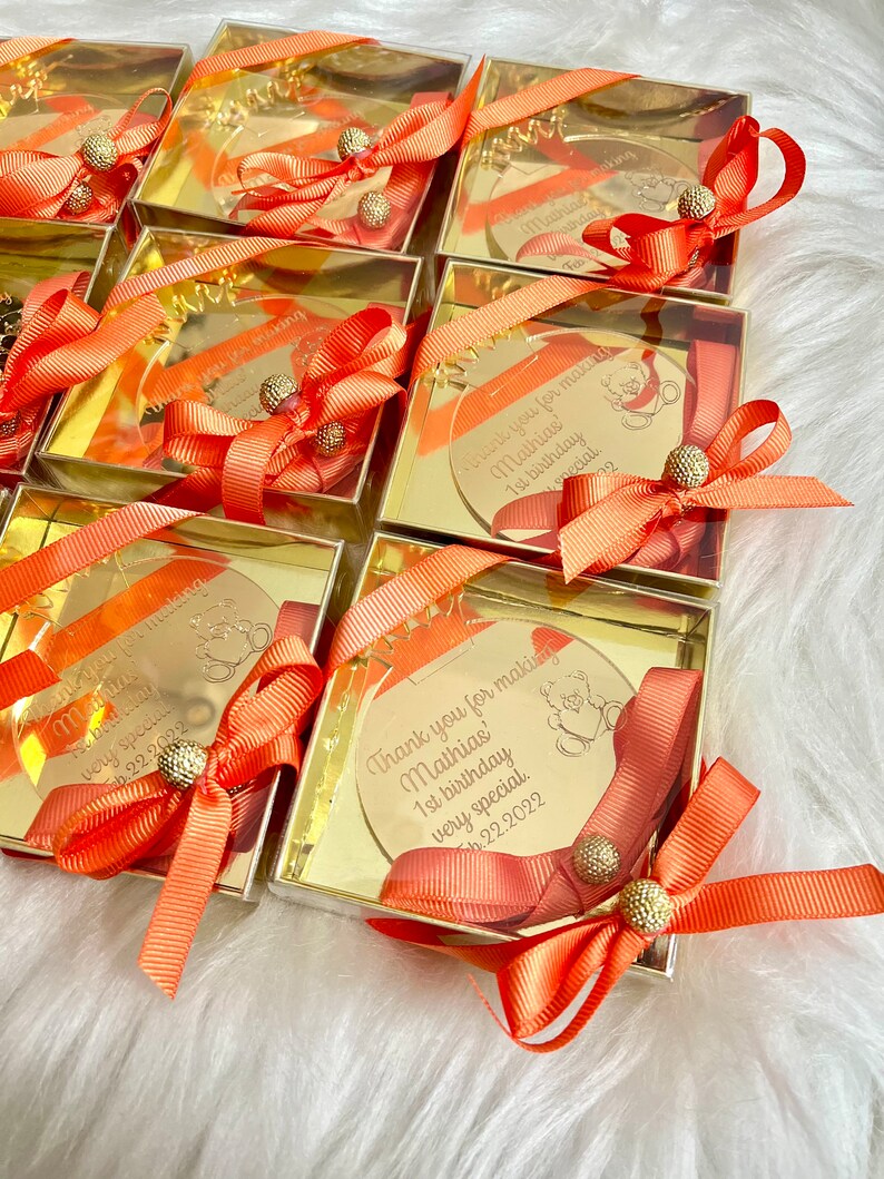 May include: Gold favor boxes with clear lids and orange ribbons. Each box contains a round, gold mirror with a personalized message that says "Thank you for making Mathias' 1st birthday very special." The date "Feb 22, 2022" is also included on the mirror.