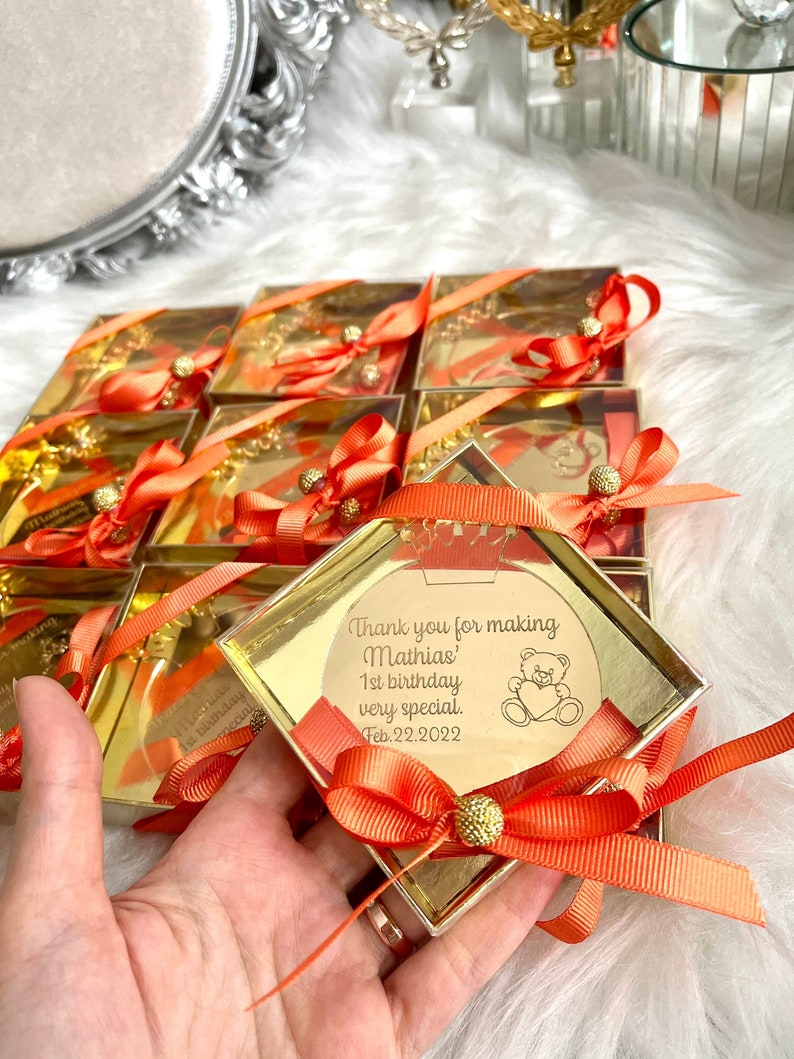 May include: Gold favor boxes with orange ribbon and a gold round charm with the text "Thank you for making Mathias' 1st birthday very special. Feb. 22. 2022" and a cartoon bear holding a heart.
