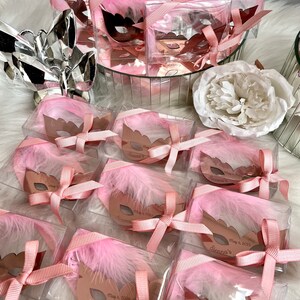 May include: Pink and gold party favours in clear boxes with a pink feather and a gold mask with the text "Sienna's" and the date "May 6, 2023".
