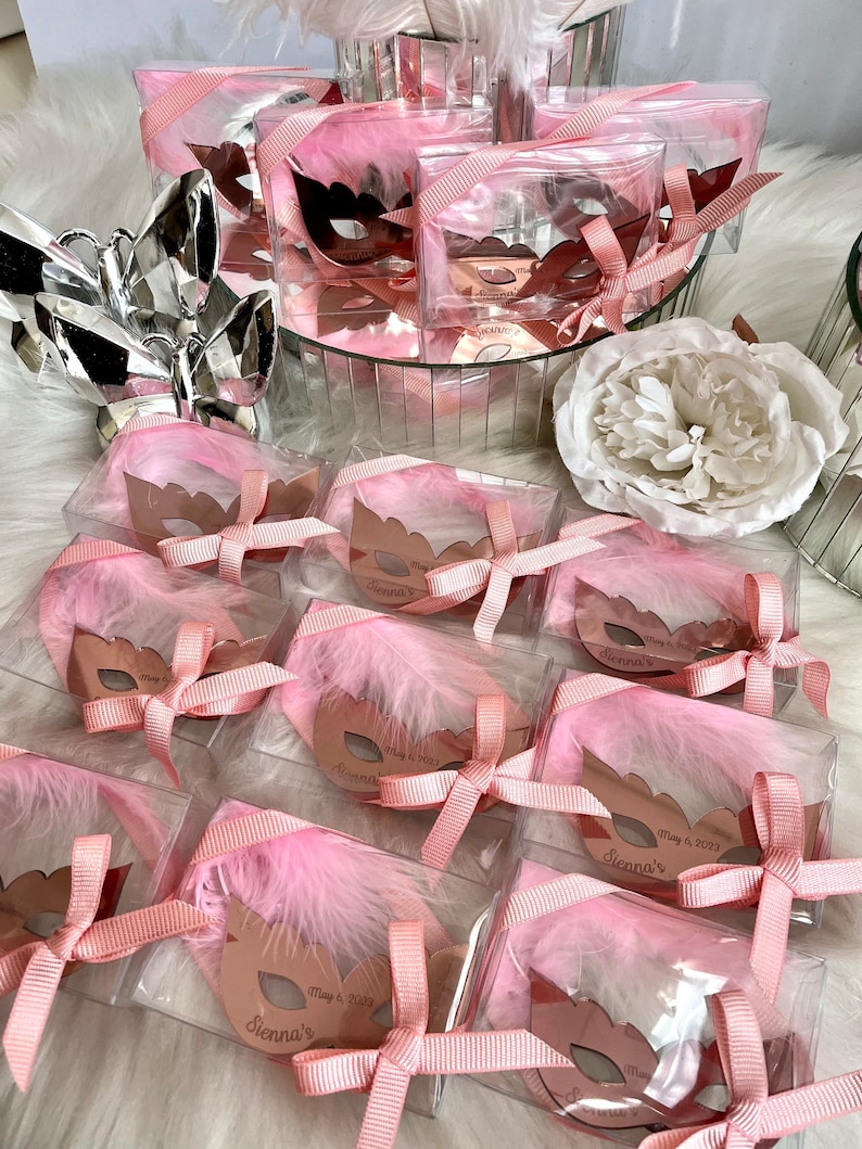 May include: Pink and gold party favours in clear boxes with a pink feather and a gold mask with the text "Sienna's" and the date "May 6, 2023".