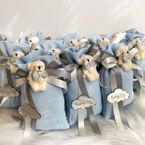 Puede incluir: Bolsas de regalo azul claro con un peque&ntilde;o osito de peluche con una pajarita. Cada bolsa tiene una etiqueta plateada con el texto "Baby CJ" en forma de nube.