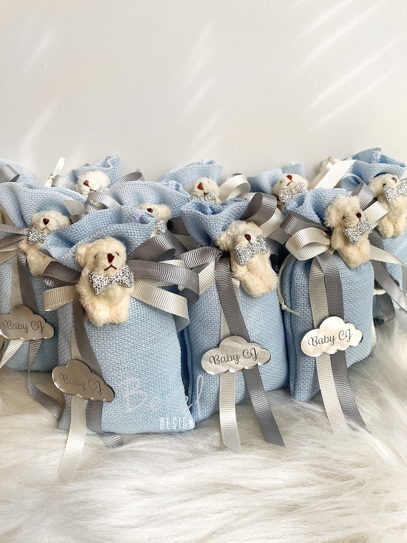Puede incluir: Bolsas de regalo azul claro con un peque&ntilde;o osito de peluche con una pajarita. Cada bolsa tiene una etiqueta plateada con el texto "Baby CJ" en forma de nube.