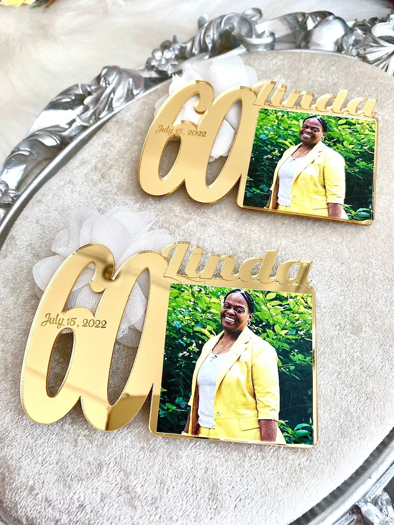 May include: Two gold mirror acrylic plaques with the number 60 and the name Linda. The plaques have a photo of a person in a yellow jacket. The text on the plaques reads "July 15, 2022".