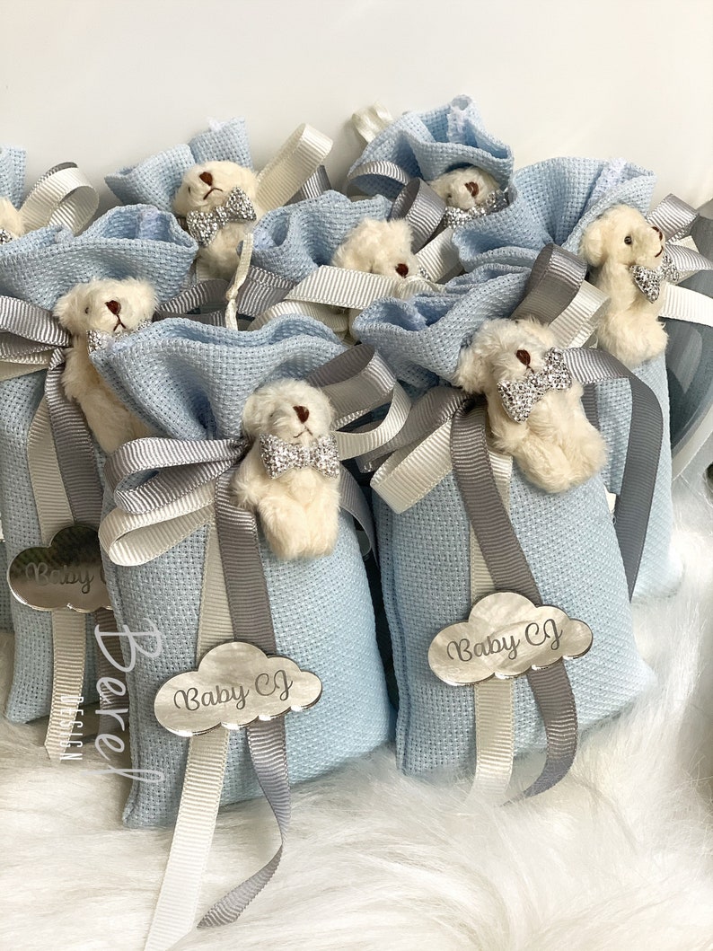 Puede incluir: Bolsas de regalo azul claro con ositos de peluche blancos que llevan pajaritas. Cada bolsa tiene una etiqueta en forma de nube plateada con el texto "Baby CJ" en ella.