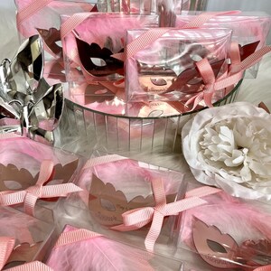 May include: Pink favour boxes with clear lids filled with white feathers and a rose gold mirror mask with the name "Sienna" and the date "May 3, 2023".