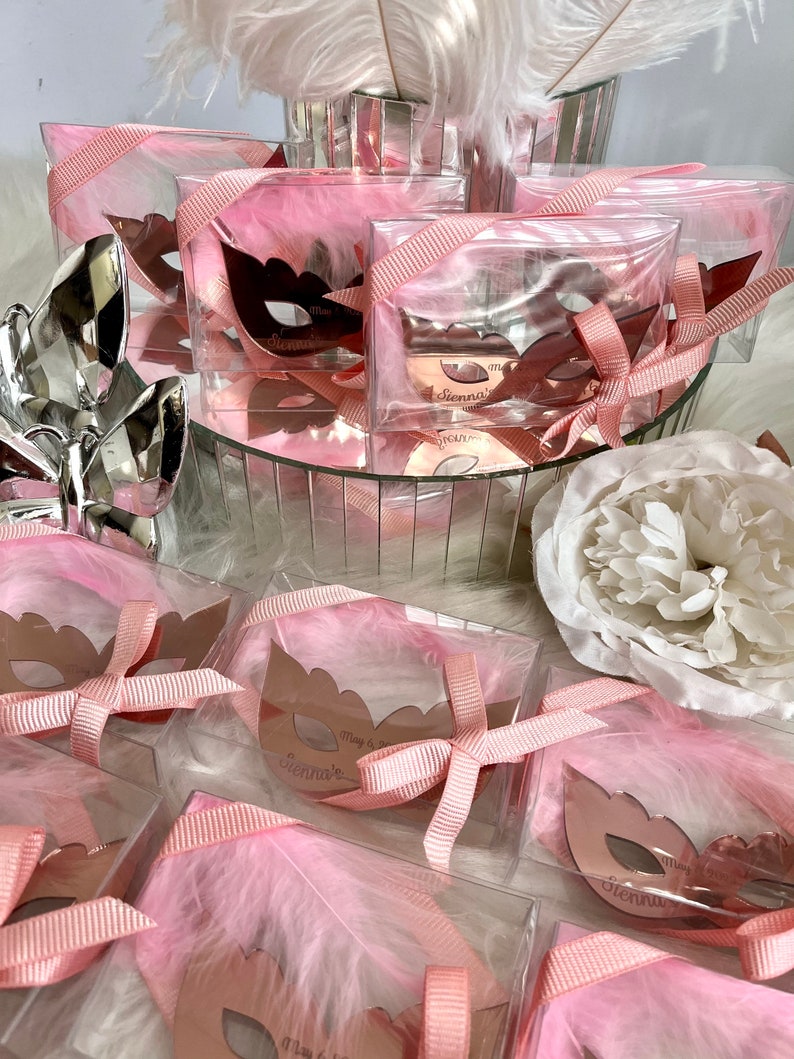 May include: Pink favour boxes with clear lids filled with white feathers and a rose gold mirror mask with the name "Sienna" and the date "May 3, 2023".
