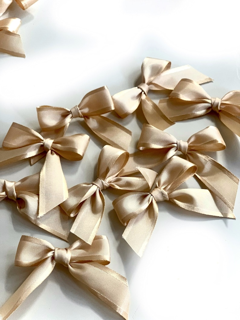 May include: A collection of ten beige satin bows, arranged in a cluster on a white surface. The bows are tied with a simple knot and have a soft, elegant look.