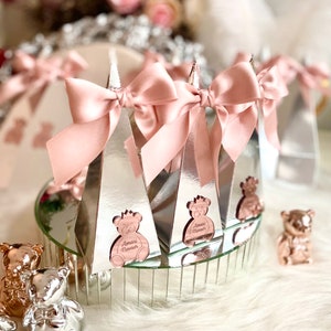 May include: Silver favour boxes with pink bows and a rose gold teddy bear design. Each box has the name "Amari Novah" printed on it.