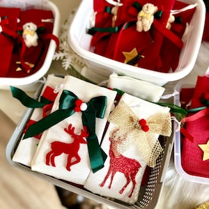 Puede incluir: Dos bolsas de regalo de tela blanca con renos rojos y lazos verdes y dorados. Las bolsas est&aacute;n en una cesta de alambre gris. Las bolsas est&aacute;n llenas de peque&ntilde;as bolsas de regalo rojas con lazos verdes y dorados.