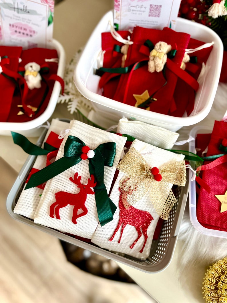 Puede incluir: Dos bolsas de regalo de tela blanca con renos rojos y lazos verdes y dorados. Las bolsas est&aacute;n en una cesta de alambre gris. Las bolsas est&aacute;n llenas de peque&ntilde;as bolsas de regalo rojas con lazos verdes y dorados.
