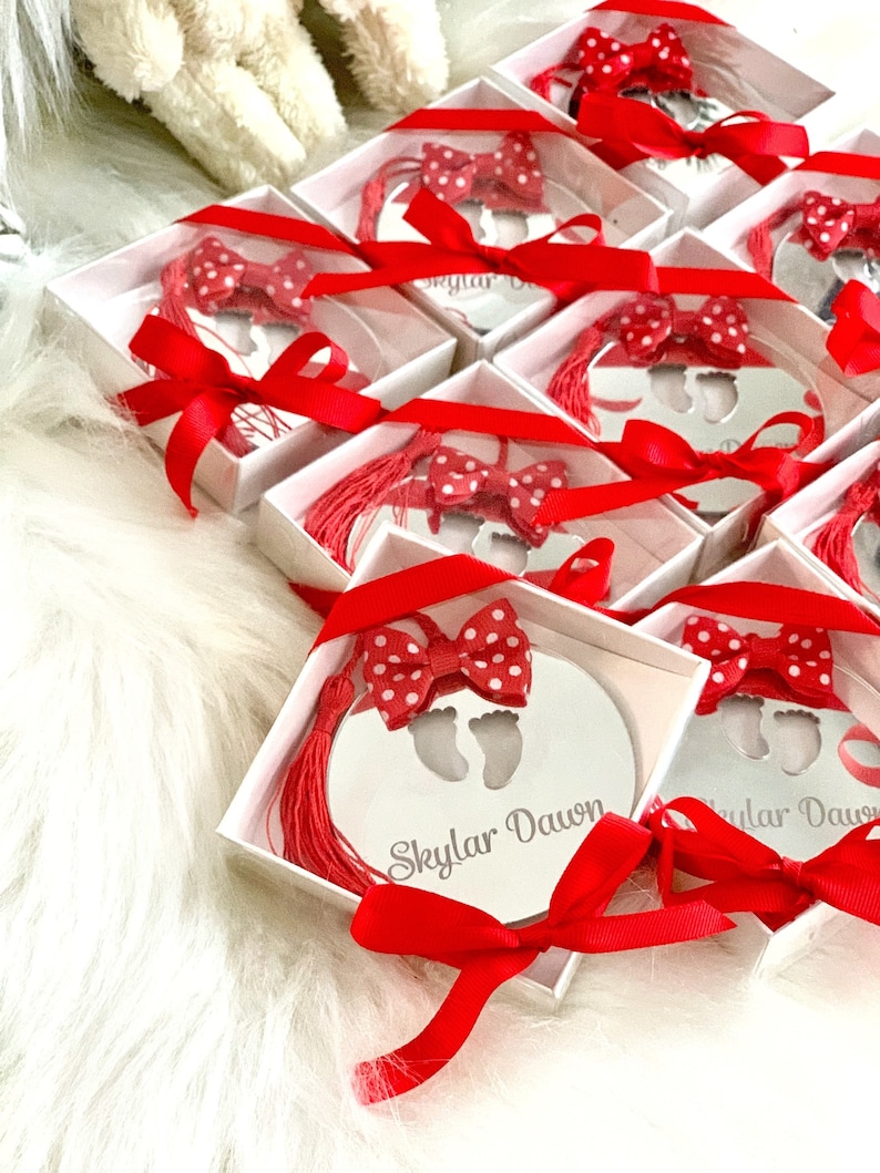 Op de afbeelding: Een set kleine, witte geschenkdozen met rood lint en een rode strik met witte stippen. Elke doos bevat een rond zilveren ornament met een baby-voetafdruk ontwerp en de naam "Skylar Dawn" gegraveerd erop.