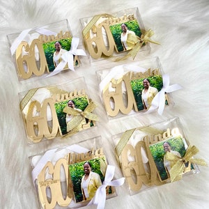 May include: Six clear boxes with gold glitter and white ribbon bows contain gold mirrored acrylic ornaments with the number 60 and the name Linda. Each ornament also has a small photo of a woman in a yellow shirt. The boxes are on a white fluffy surface.