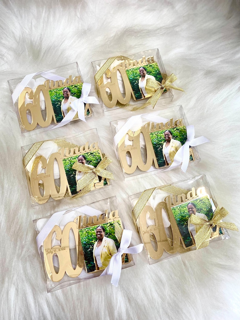 May include: Six clear boxes with gold glitter and white ribbon bows contain gold mirrored acrylic ornaments with the number 60 and the name Linda. Each ornament also has a small photo of a woman in a yellow shirt. The boxes are on a white fluffy surface.