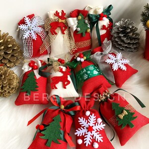 Puede incluir: Una colecci&oacute;n de 11 peque&ntilde;as bolsas de regalo de tela roja y blanca con diversas decoraciones de tem&aacute;tica navide&ntilde;a, incluyendo copos de nieve, renos, &aacute;rboles de Navidad y lazos. Las bolsas est&aacute;n dispuestas sobre una superficie blanca y esponjosa.