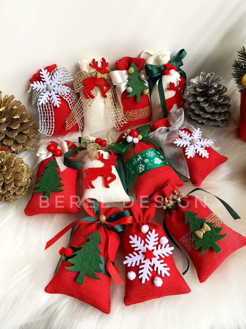 Puede incluir: Una colecci&oacute;n de 11 peque&ntilde;as bolsas de regalo de tela roja y blanca con diversas decoraciones de tem&aacute;tica navide&ntilde;a, incluyendo copos de nieve, renos, &aacute;rboles de Navidad y lazos. Las bolsas est&aacute;n dispuestas sobre una superficie blanca y esponjosa.