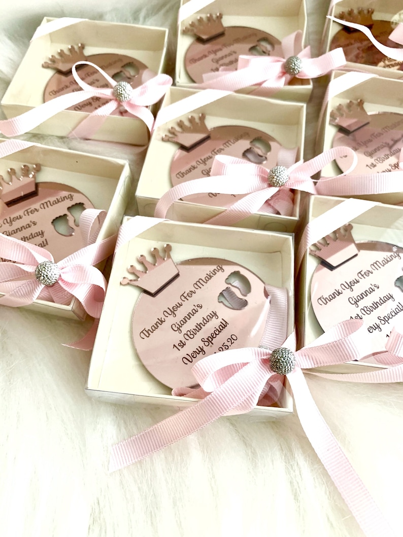 May include: Pink and white favor boxes with a rose gold crown and a round mirror with the text "Thank You For Making Gianna's 1st Birthday Very Special!"