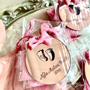 Op de afbeelding: Roze en gouden babyshower-souvenir met een roze strik en kwast. Het souvenir is een ronde spiegel met de tekst "Nyla Meilani Brazil 2021" en een paar babyvoetjes.