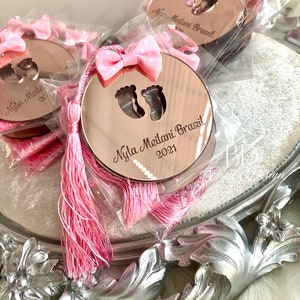 Op de afbeelding: Roze en gouden babyshower-cadeautjes met een roze kwast en een ronde spiegel met de tekst "Nyla Meilani Brazil 2021" en een paar babyvoetjes.