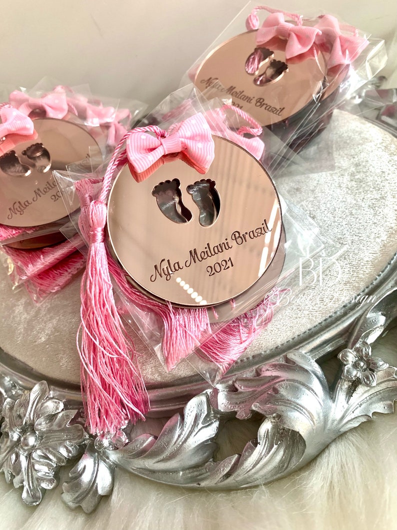Op de afbeelding: Roze en gouden babyshower-cadeautjes met een roze kwast en een ronde spiegel met de tekst "Nyla Meilani Brazil 2021" en een paar babyvoetjes.